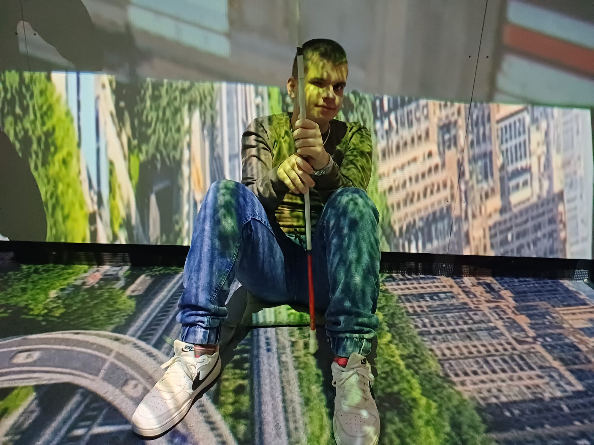 Mladić sjedi na podu u prostoriji s projekcijom grada po podu i zidovima, u rukama drži bijeli štap i okružen je svjetlima projekcije.