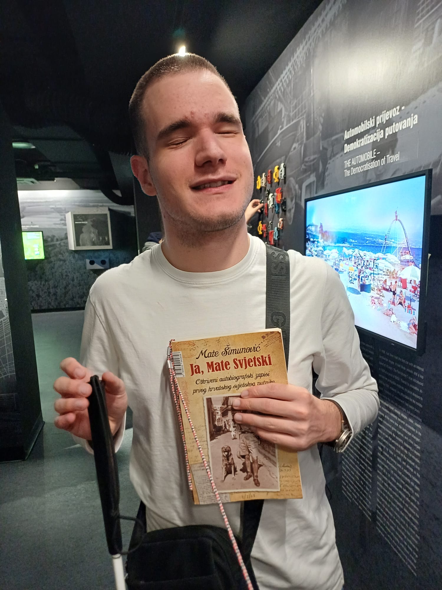 Mladić u bijeloj majici stoji u muzeju s bijelim štapom u jednoj ruci, a u drugoj drži knjigu „Ja, Mate Svjetski“, lice mu je blago nasmiješeno.
