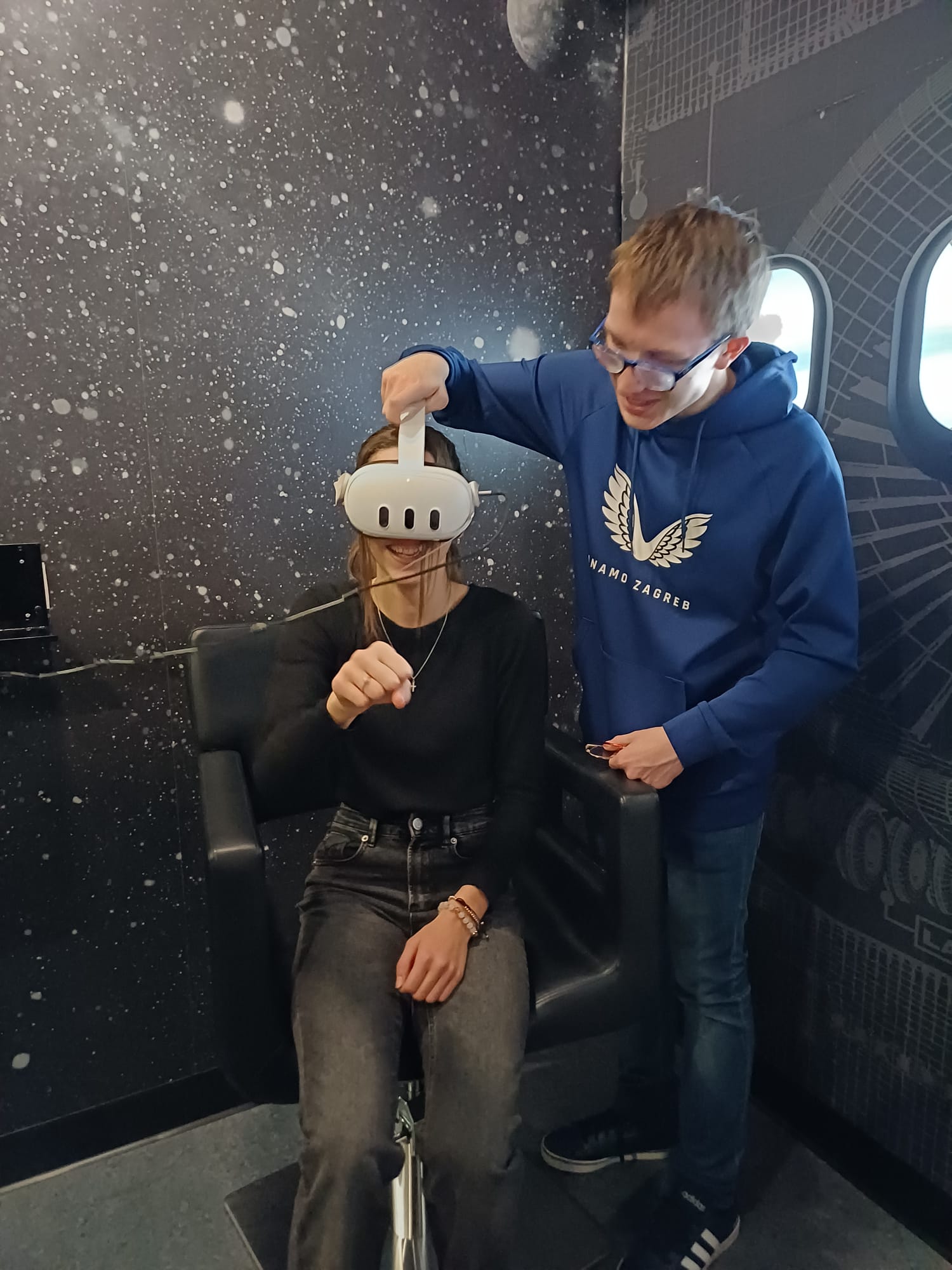 Djevojka u crnoj majici i trapericama sjedi u tamnoj stolici u prostoru sa zvjezdastom pozadinom, na glavi ima VR naočale, a pored nje stoji mladić u plavoj trenirci i namješta joj naočale.