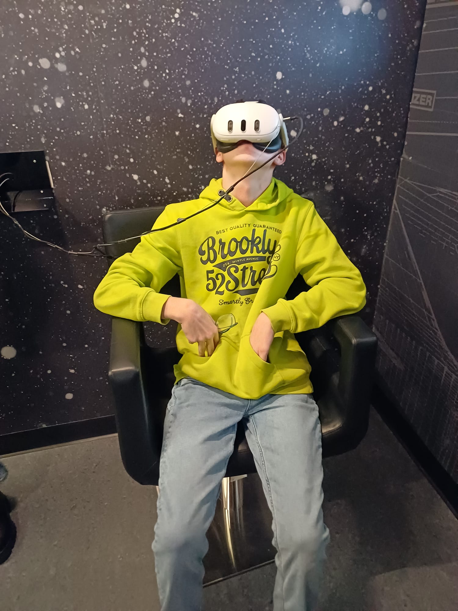 Učenik u žutoj hoodici i trapericama sjedi zavaljen u tamnoj stolici, na glavi ima VR naočale, a ruke su mu opušteno u džepovima hoodice; pozadina je tamna sa svjetlucavim točkicama nalik zvijezdama.