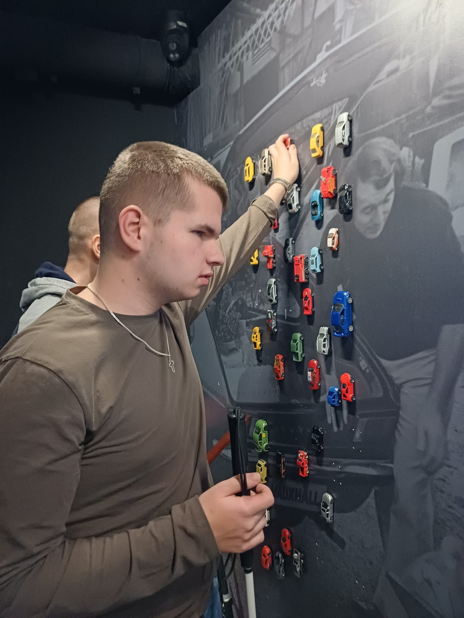 Mladić u smeđoj majici stoji ispred zida prekrivenog malim šarenim autićima i dotiče jedan model rukom, u drugoj ruci drži sklopljeni bijeli štap.