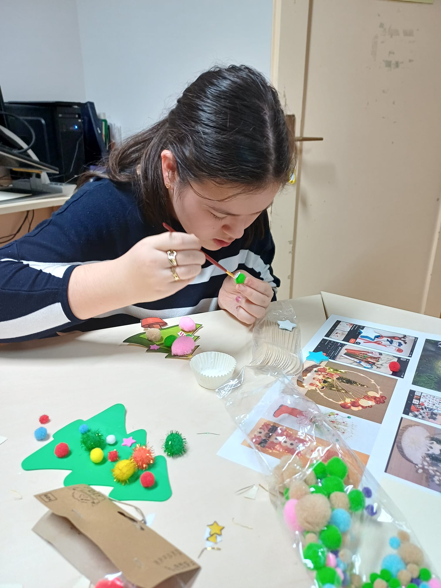 Korisnica sjedi za stolom u učionici i izrađuje božićnu jelku od zelenog kartona ukrašenu pomponima i zvjezdicama.