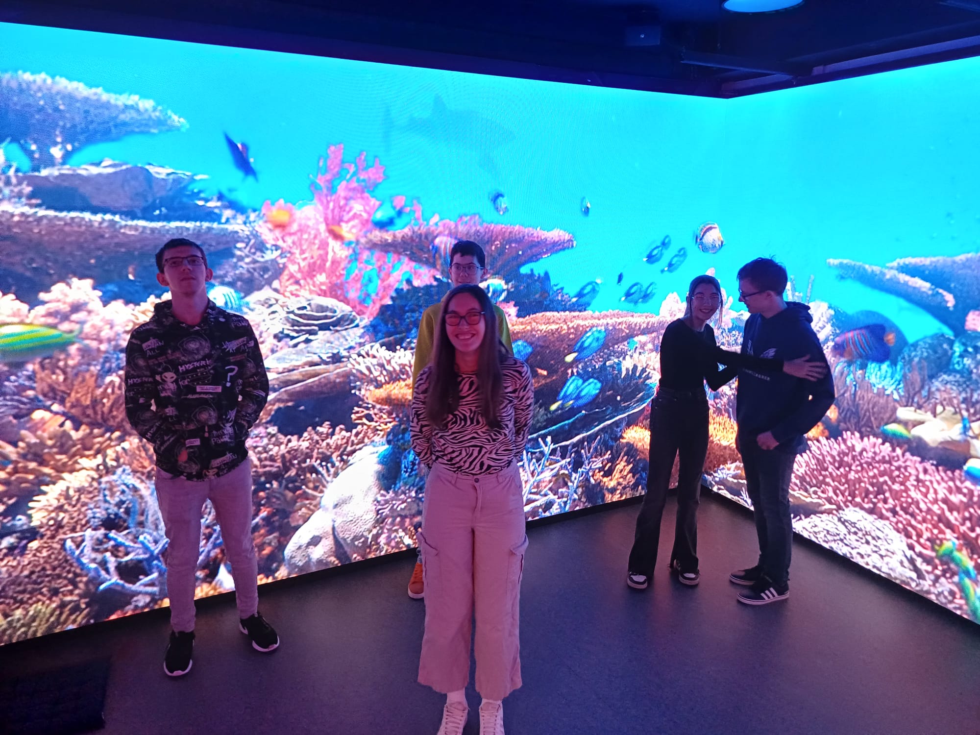 Nekoliko učenika stoji u prostoriji s velikim ekranom na kojem se prikazuje šarena podvodna scena s koraljima i ribama; jedan učenik stoji bliže kameri, ostali su raspoređeni uz rubove prostora.