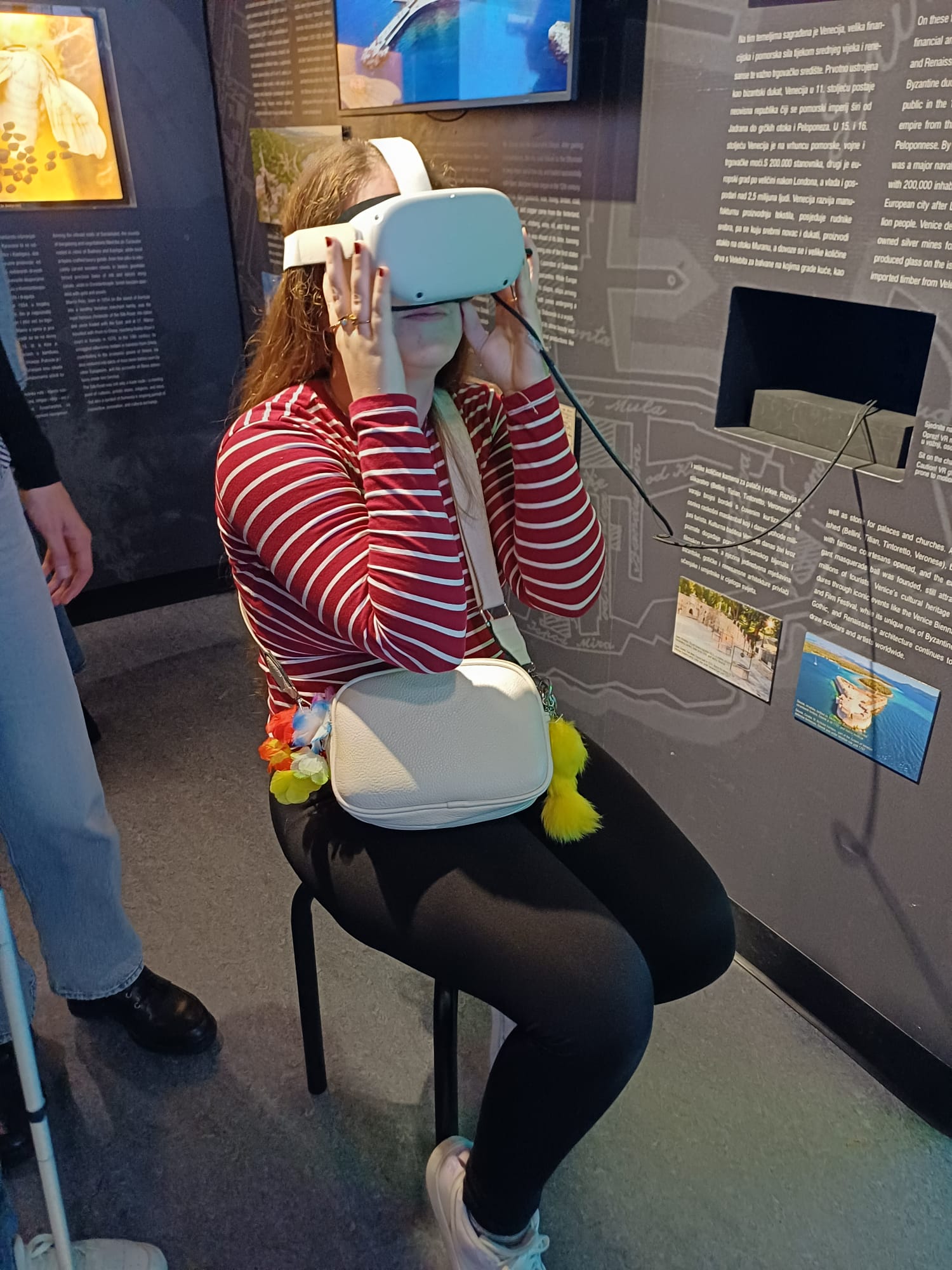 Djevojka s dugom kosom sjedi na stolcu u muzeju, nosi VR naočale i drži ih rukama na licu; u pozadini su informativni paneli i fotografije putovanja.