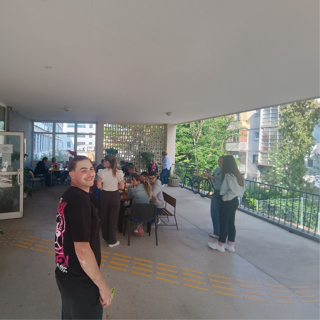 Učenik Eron se smiješi dok željno iščekuje pečeno meso, u pozadini grupa učenika za stolom.