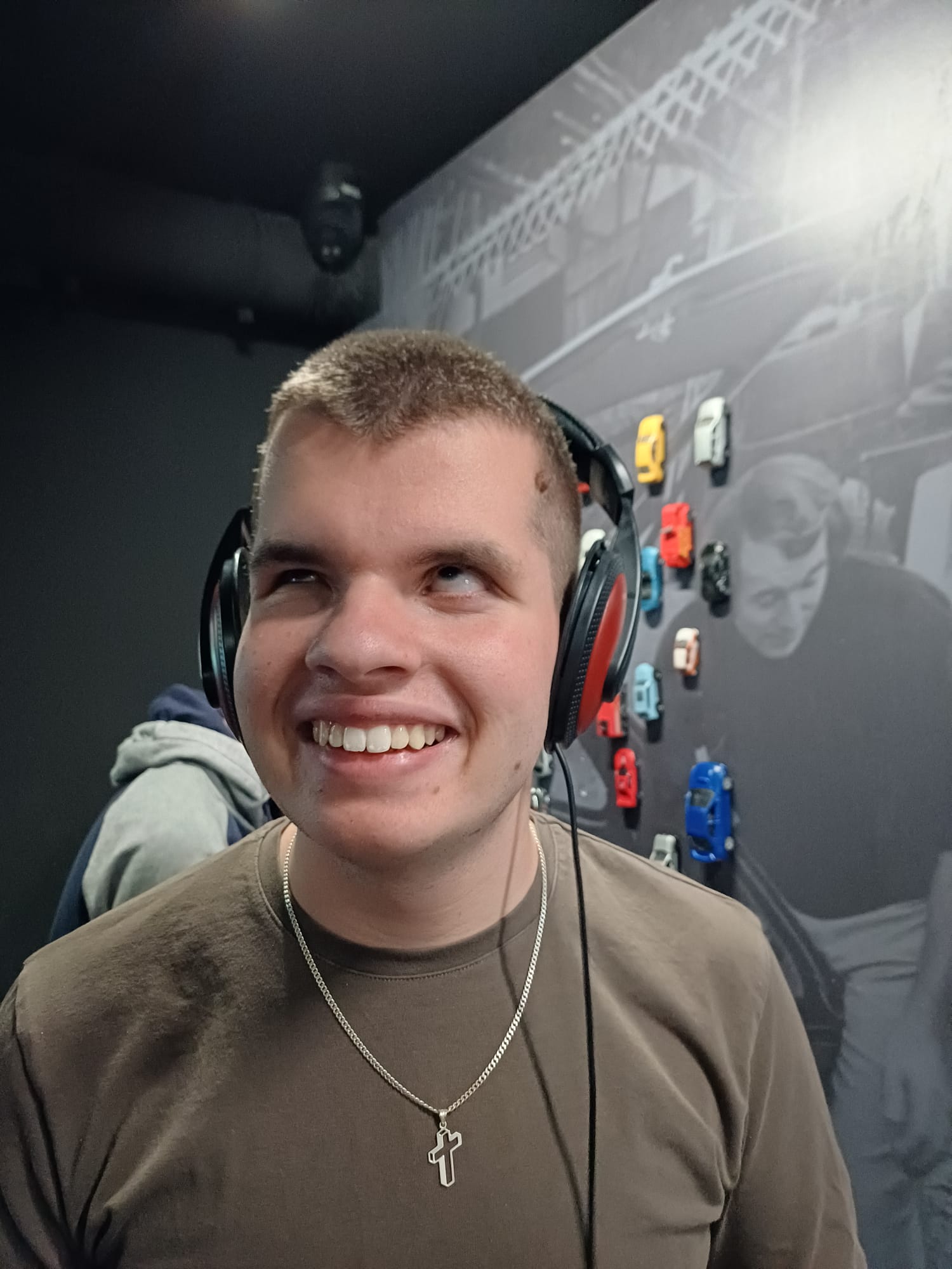 Mladić kratke kose u smeđoj majici stoji ispred zida s malim modelima automobila, nasmijan je i nosi velike slušalice preko ušiju.